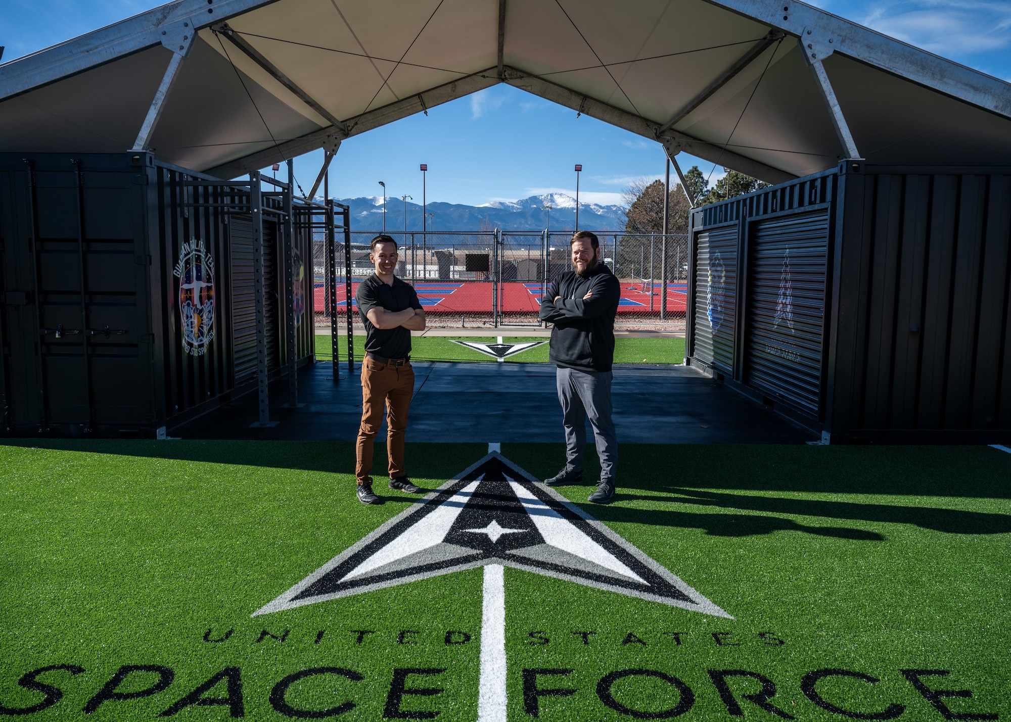 Two men stand in front of workout equipment outside.
