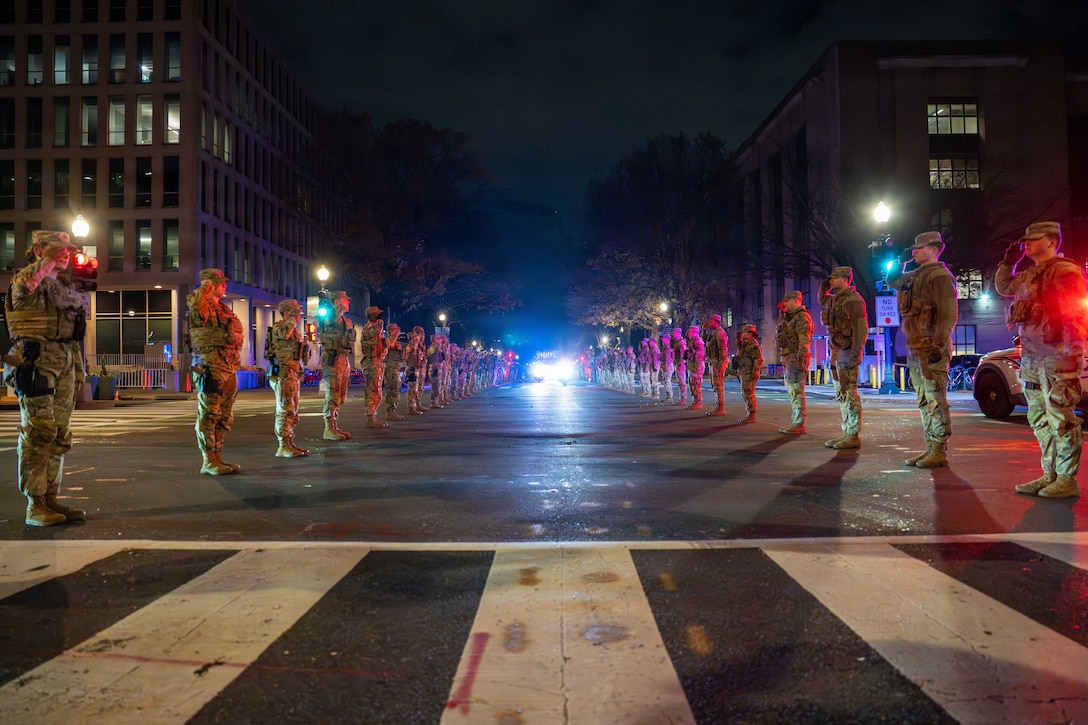 Service members wearing camouflage uniforms and facing each other in two lines while standing on a dark street salute as a vehicle with headlights on moves forward between the facing service members.