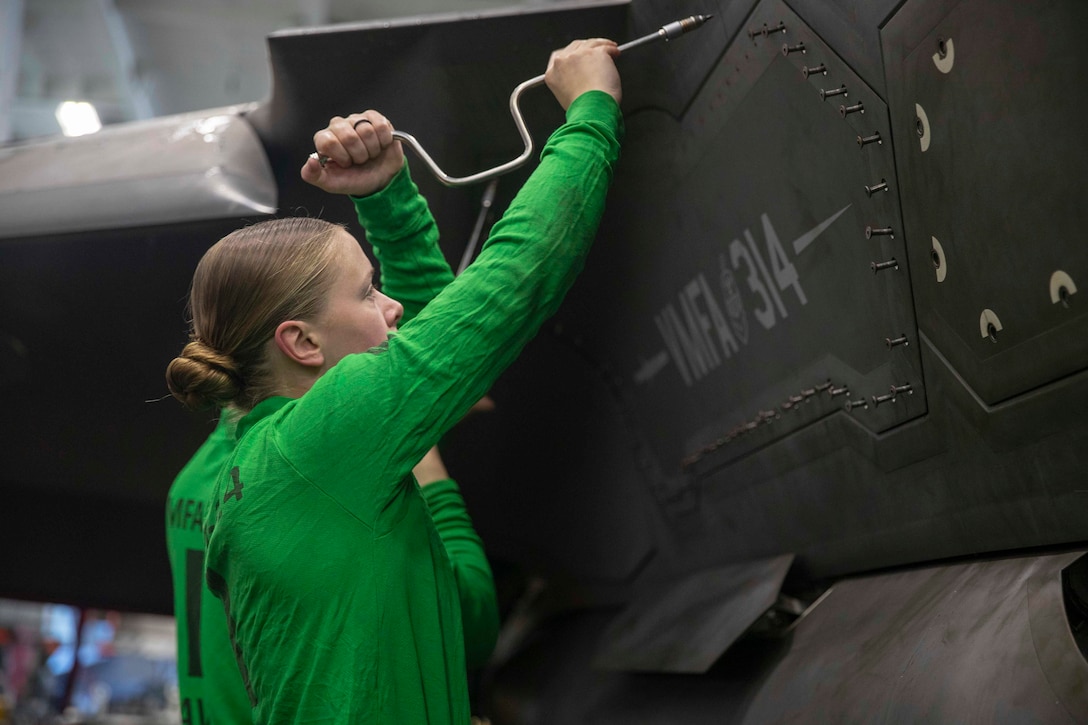 A Marine uses a handheld tool to adjust screws on the panel of an aircraft indoors, with another person in the background.