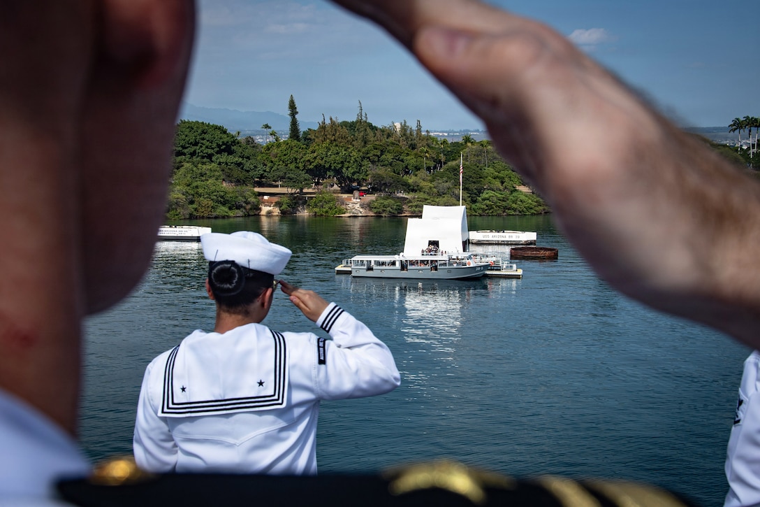 Sailors aboard a ship salute as they pass a memorial in a body of water during daytime, with trees in the background.