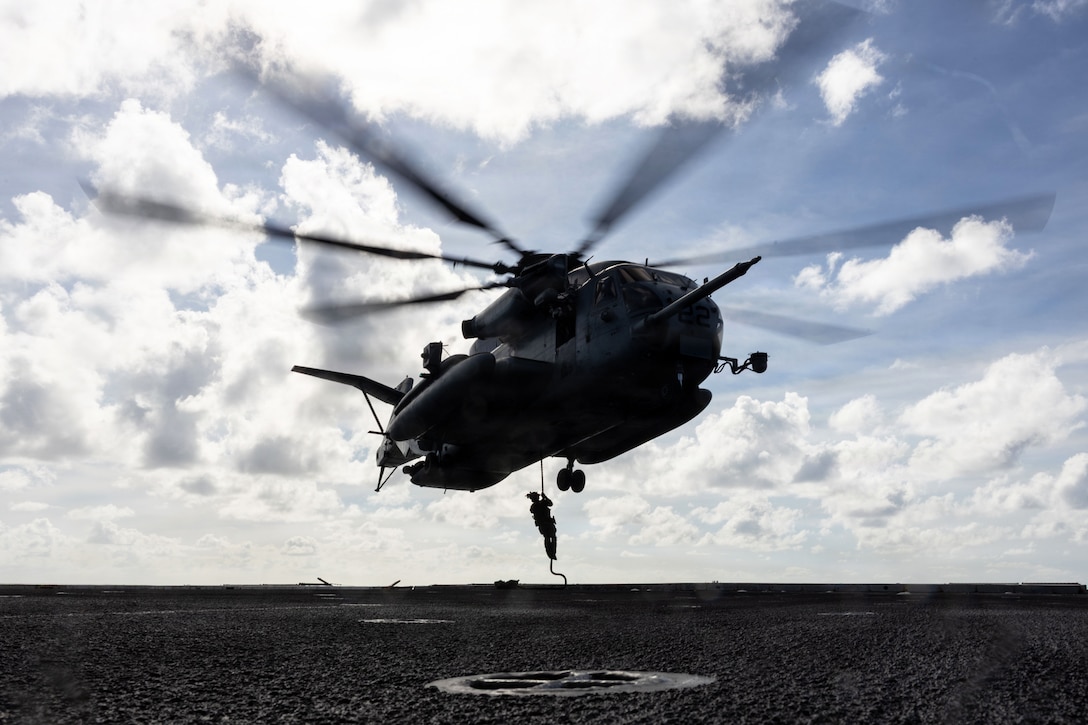 A Marine descends a rope attached to a helicopter hovering over an object floating in a body of water on a cloudy day.