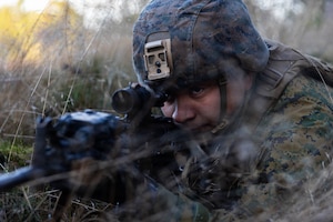 A Marine in combat gear and lying in a grassy area looks through the scope of a rifle during daytime.