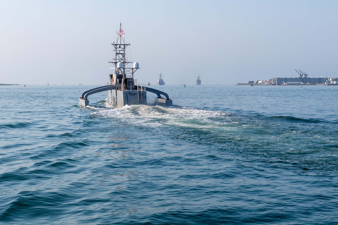 An autonomous vessel moves in a large body of water during daytime, with other boats and buildings on land in the distance.