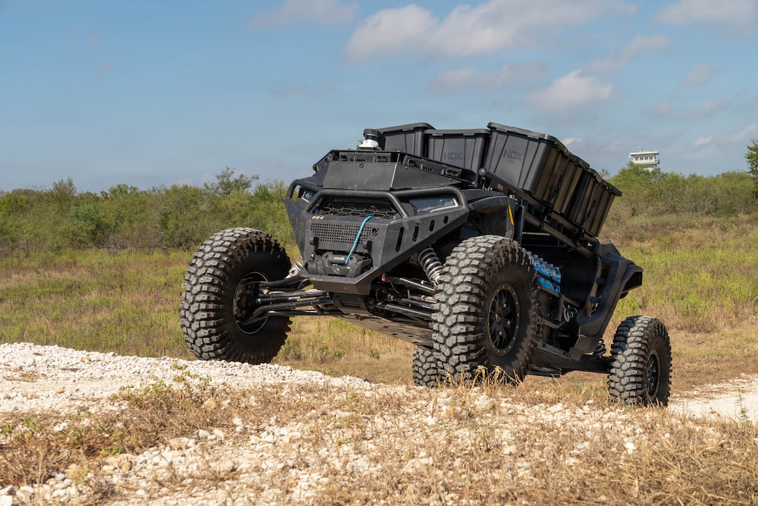 An unmanned vehicle moves on a rocky, sandy road during daytime, with green vegetation and a building in the background.