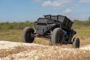 An autonomous unmanned vehicle treks down a dirt path in a grassy field with blue sky and clouds.