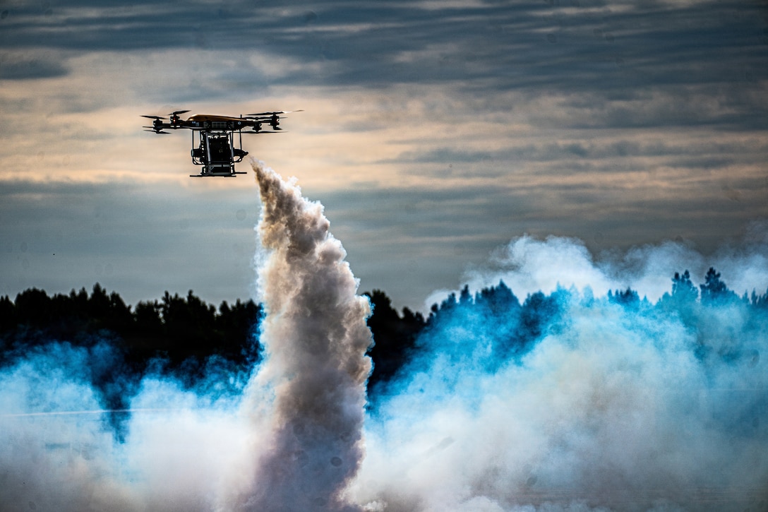 An unmanned aircraft blows a smoky substance billowing toward the ground, with trees and clouds in a dusk-like sky.