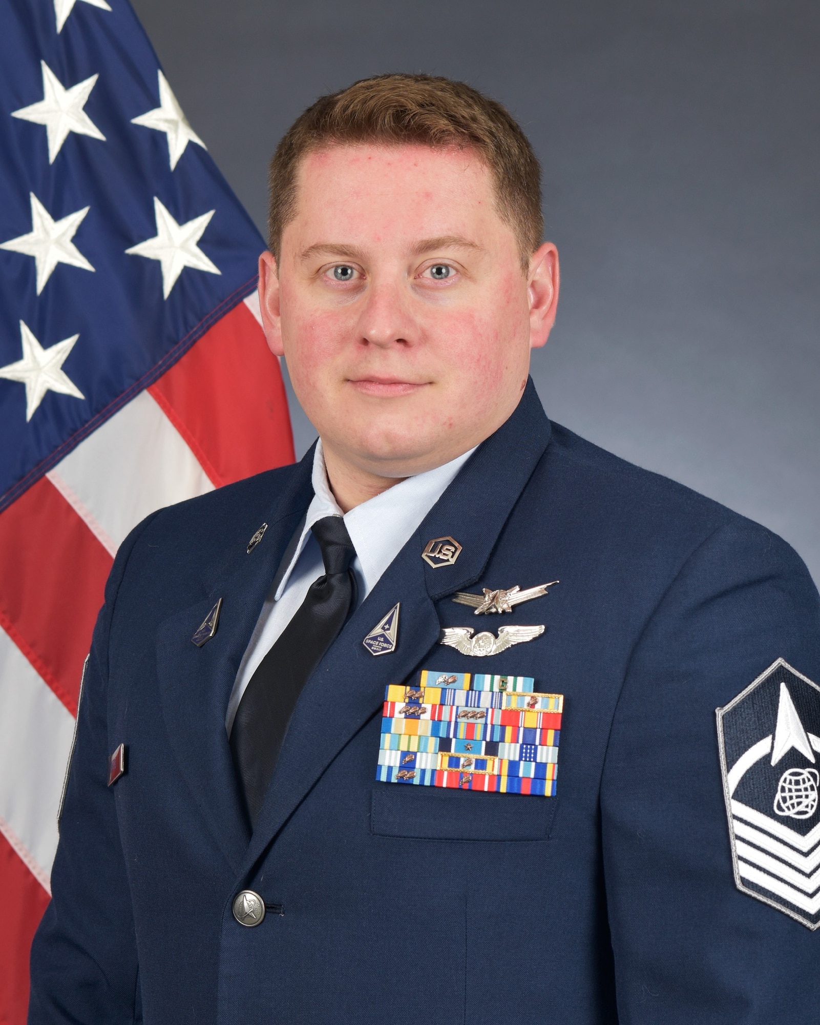 Master Sgt. Adam Spittle poses for an official photo wearing the Air Force standard blues dress uniform in front of an American flag.