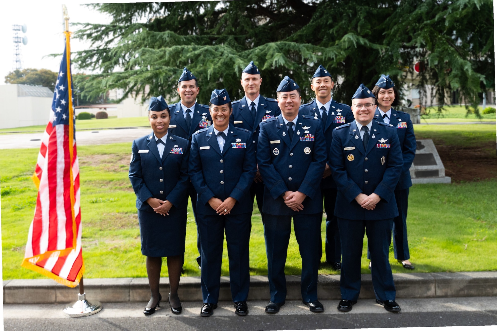 A group of eight individuals in the U.S. Space Forces Japan team poses for an official photo wearing the Air Force standard blues dress uniform outside beside an American flag.