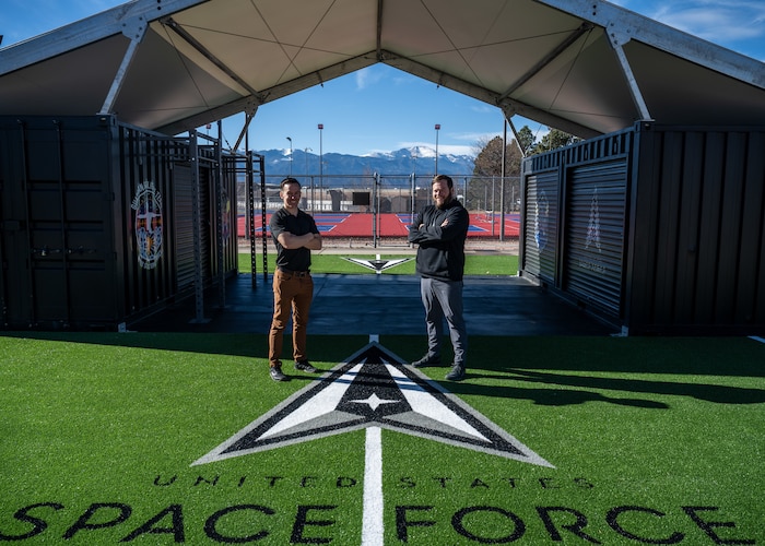 Two men stand in front of workout equipment outside.