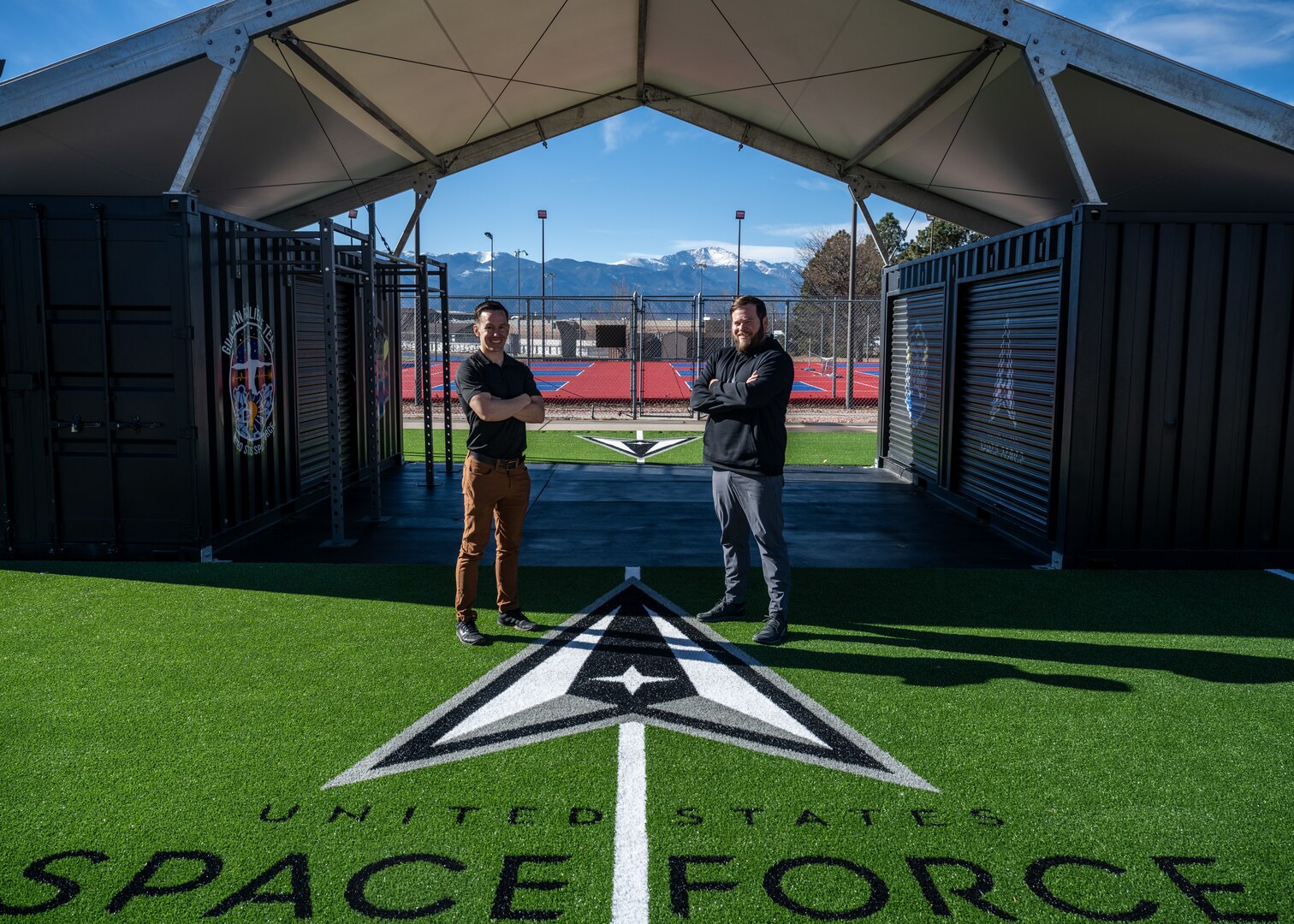 Two men stand in front of workout equipment outside.