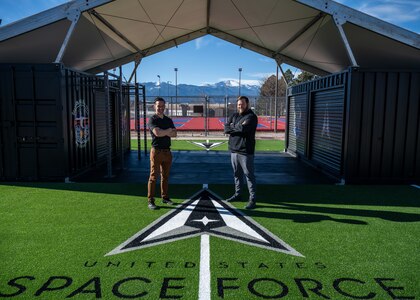 Two men stand in front of workout equipment outside.