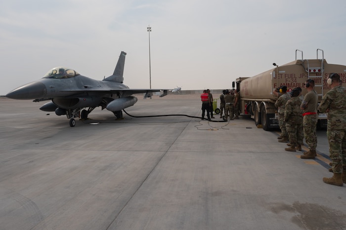 An F-16 Fighting Falcon aircraft is parked on the left side. A hose runs from it to a refueling truck parked on the right side. A dozen Airmen stand along the refueling truck watching.