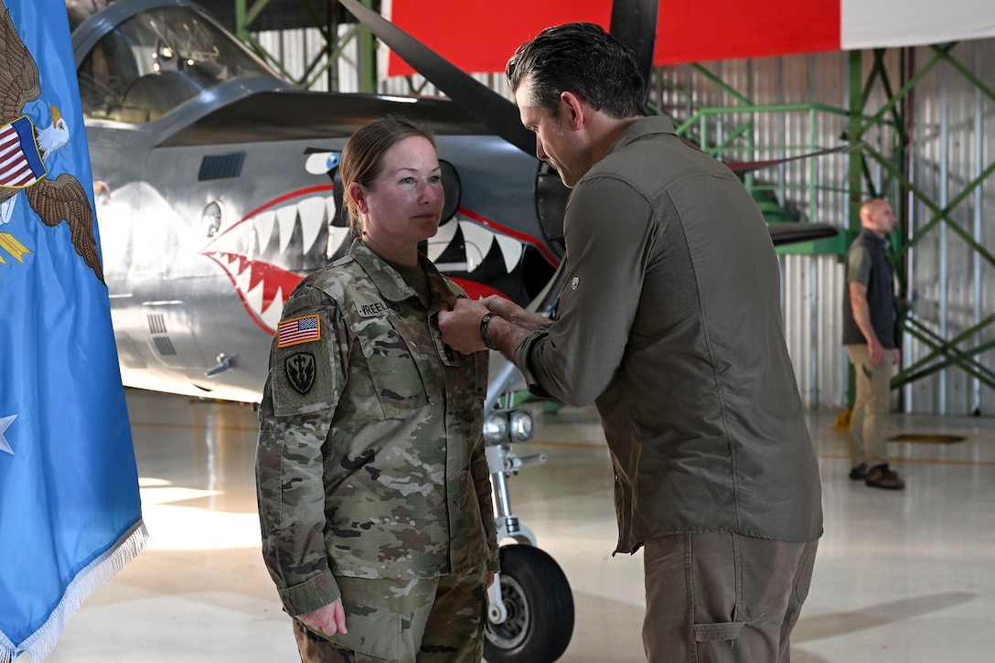 A man in civilian attire pins a medal on a woman in a camouflage military uniform inside an aircraft hangar. The Secretary of War flag is next to the woman, and the nose of a fighter aircraft is in the background.