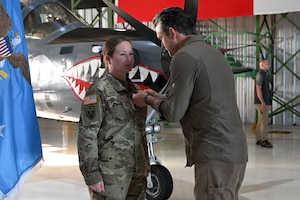 A man in civilian attire pins a medal on a woman in a camouflage military uniform inside an aircraft hangar. The Secretary of War flag is next to the woman, and the nose of a fighter aircraft is in the background.