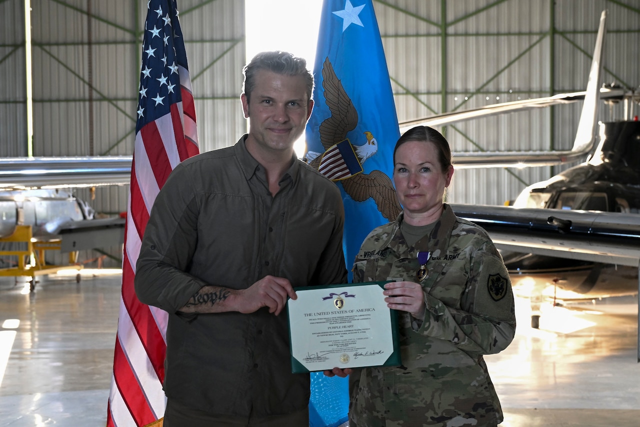 A man in civilian attire and a woman in a camouflage military uniform stand inside an aircraft hangar holding an award certificate. The American flag and the Secretary of War flag are behind them