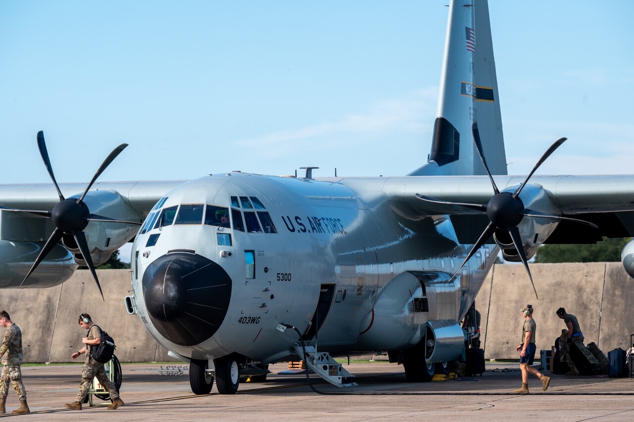Several people in military camouflage uniforms walk around a large gray military aircraft sitting on a tarmac.