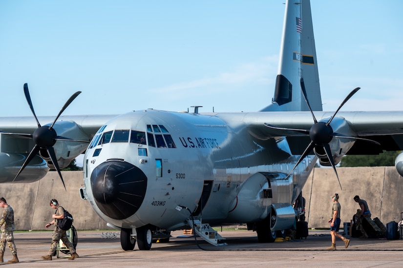 Several people in military camouflage uniforms walk around a large gray military aircraft sitting on a tarmac.