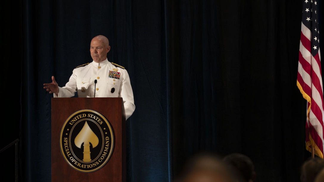 A man in a dress military uniform stands behind a lectern speaking while gesturing with his hand. A seal on the lectern shows a metallic spear tip encircled by the text "United States Special Operations Command."