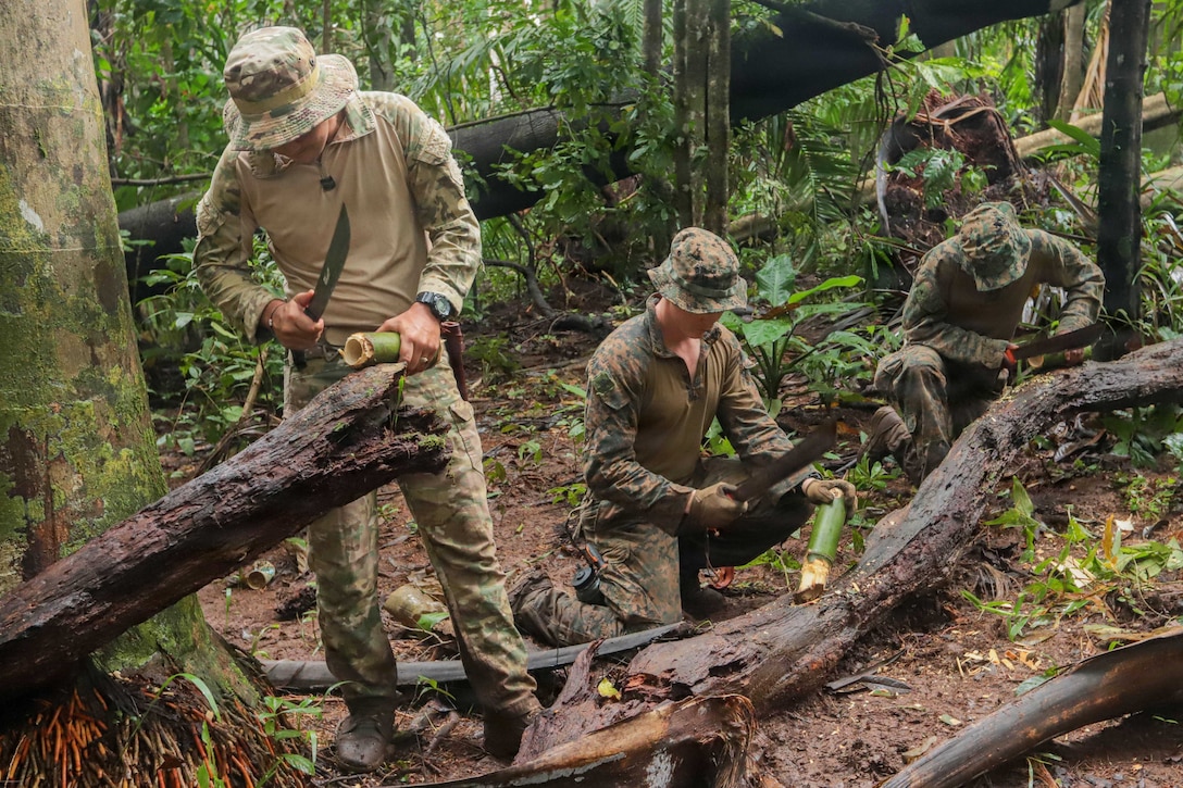 Three people wearing camouflage attire use machetes to carve pieces of bamboo during daytime in a jungle.