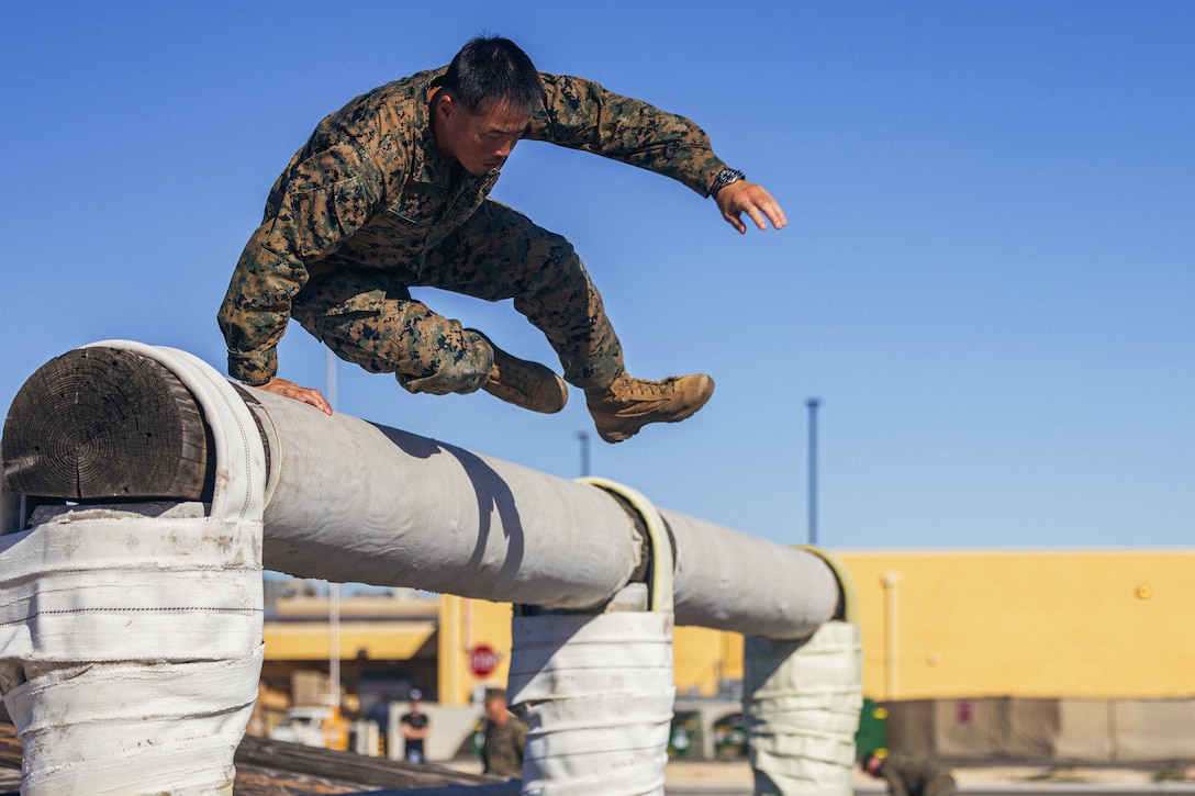A Marine in camouflage leaps over an elevated obstacle outdoors during daytime, with other Marines and a yellow building in the background.