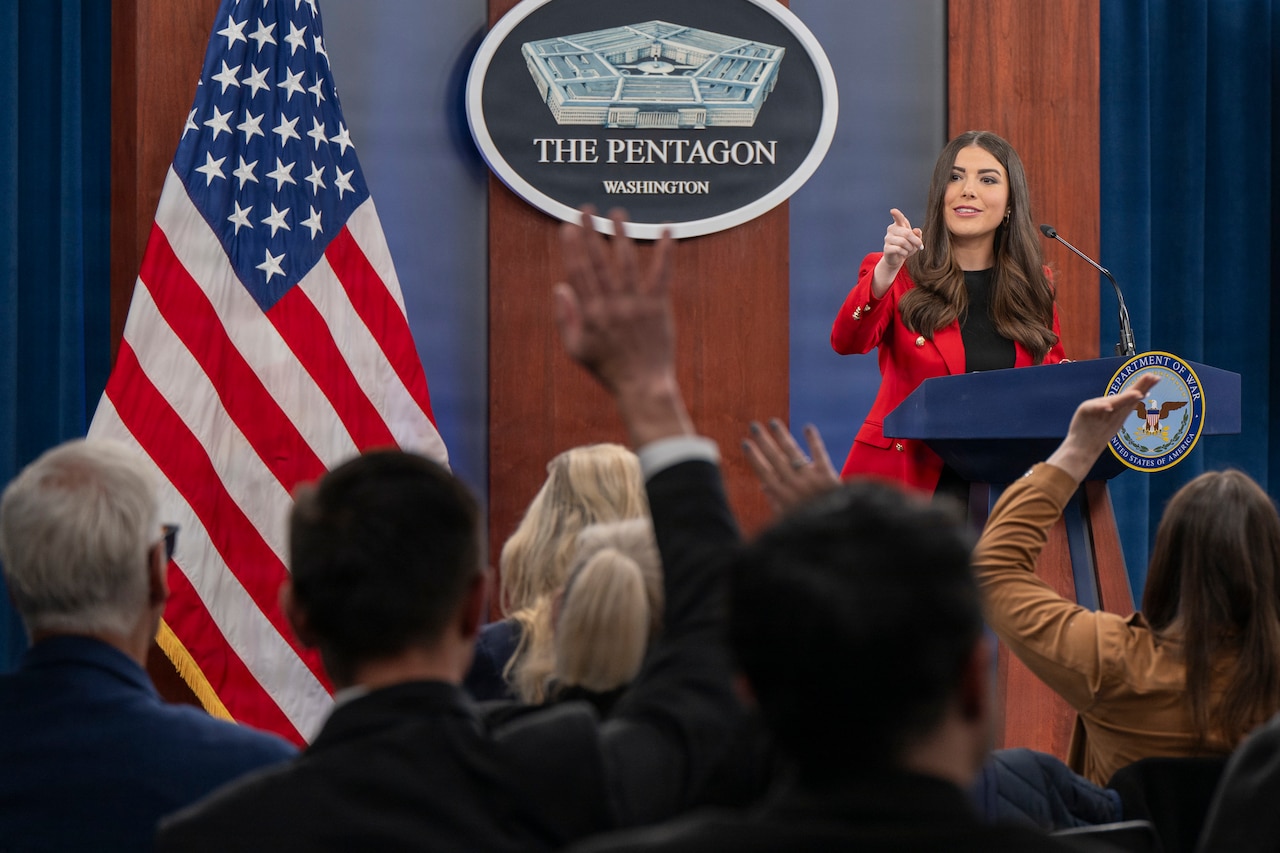 A person points from a podium as reporters raise their hands.