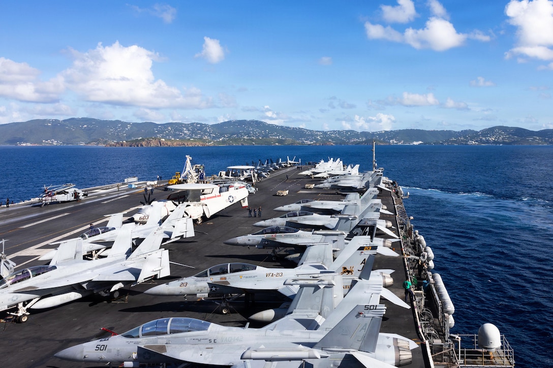 An aircraft carrier with fighter jets on its deck moves through a body of water during daytime, with hilly land in the background.