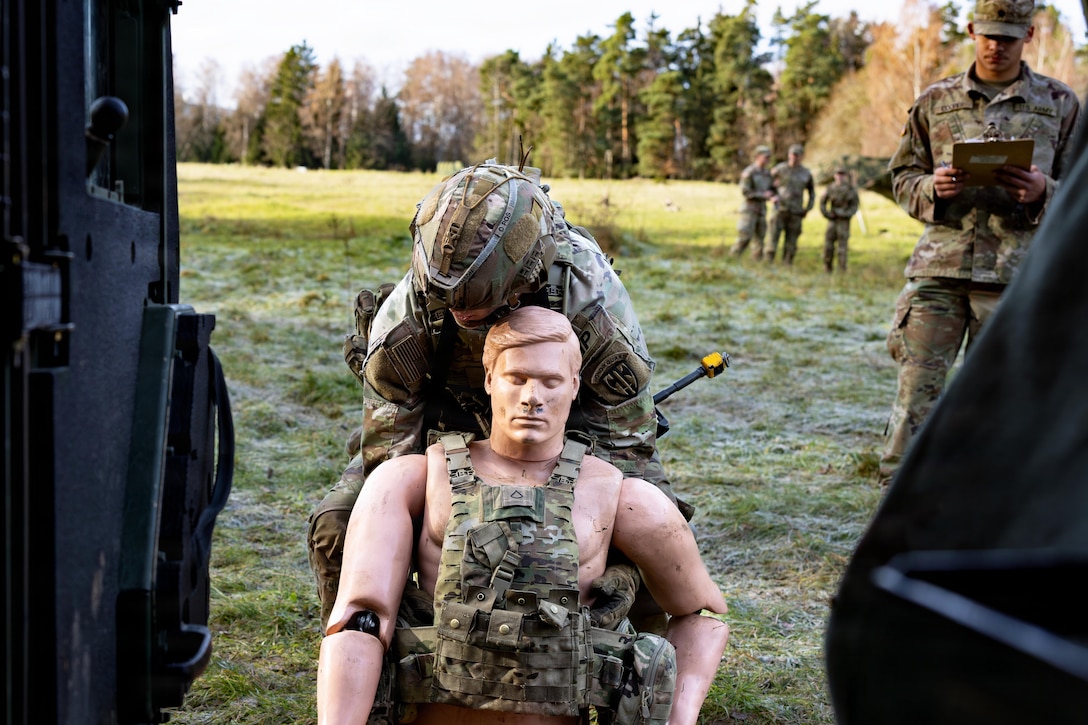 A soldier in combat gear prepares to lift a mannequin into a vehicle in a grassy area during daytime, with other soldiers and trees in the background.
