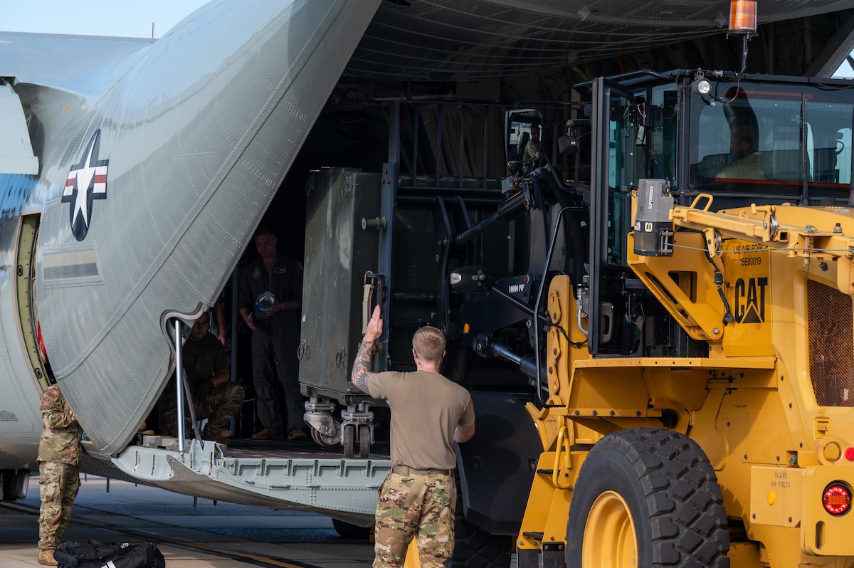 A man in a camouflage military uniform guides equipment from a forklift into the back of a military aircraft.