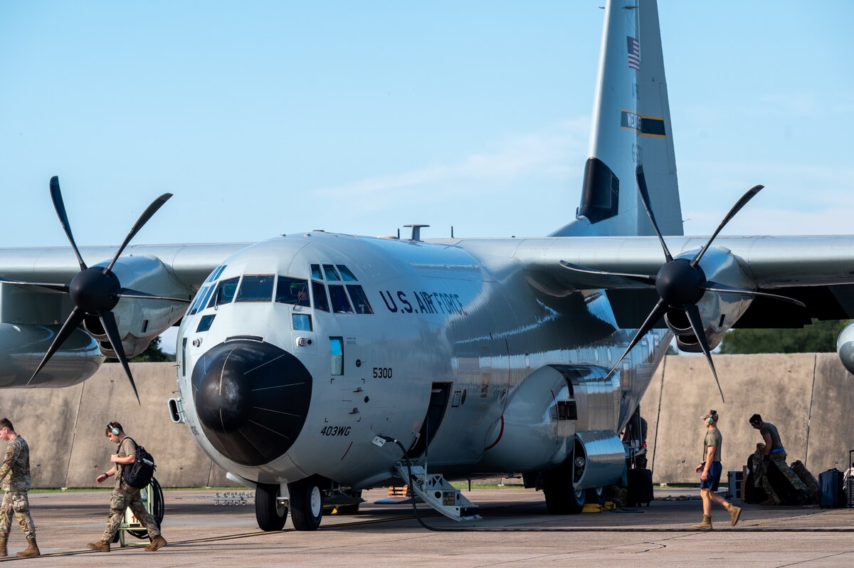 Several people in military camouflage uniforms walk around a large gray military aircraft sitting on a tarmac.