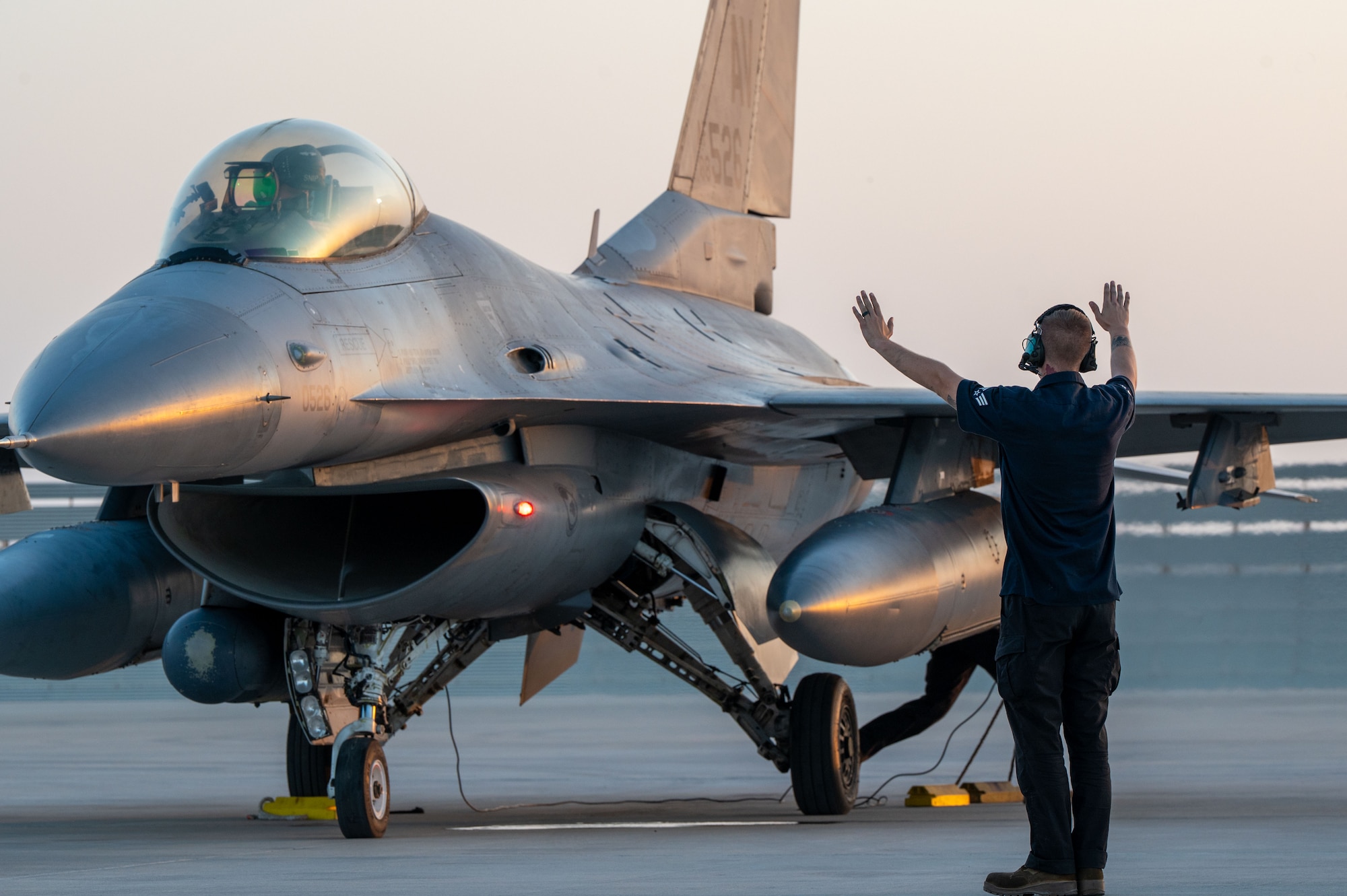 An F16 aircraft fills most of the frame. An Airman standing front, right faces the aircraft with his arms up, palms facing the aircraft.