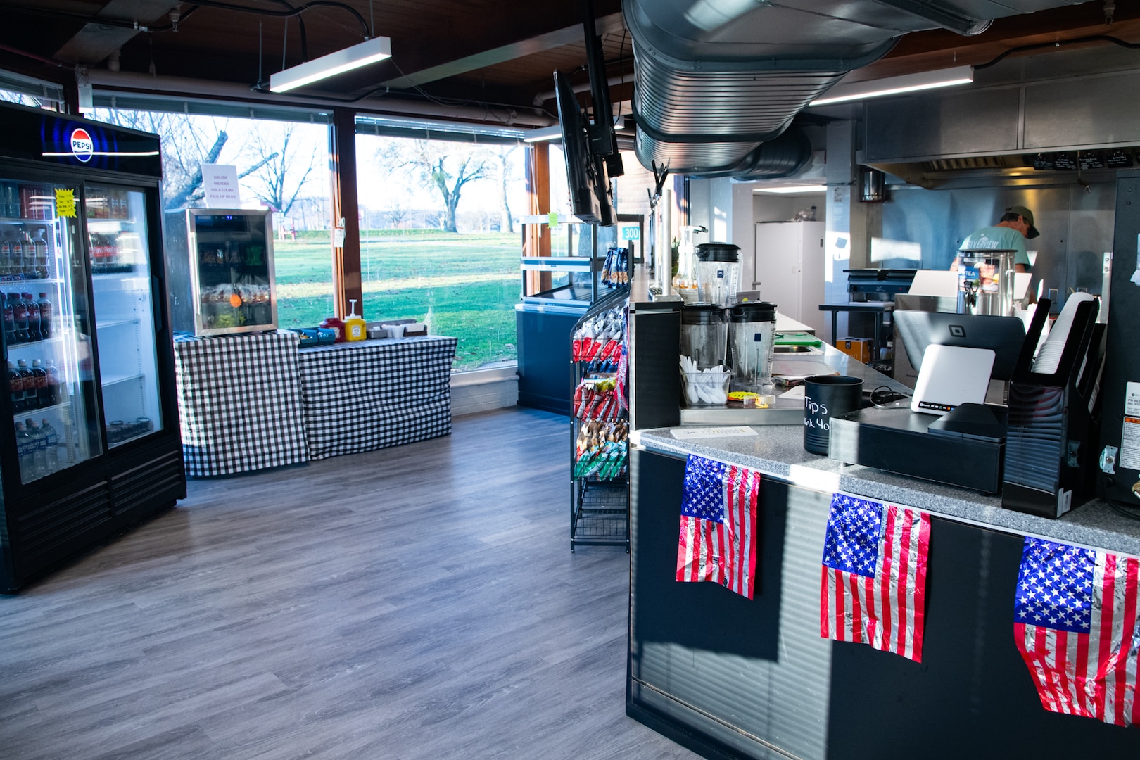 An interior of a restaurant ordering counter. There are large windows to the outdoors on the left and a counter with American flag pennants on the right.