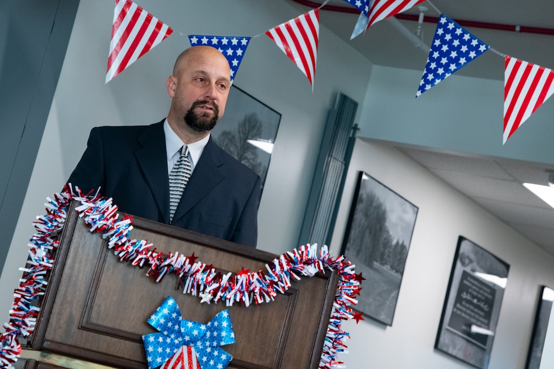 A man wearing a suit stands indoors behind a lectern speaking. There are American flag pennants behind him and red white and blue garland on the lectern.