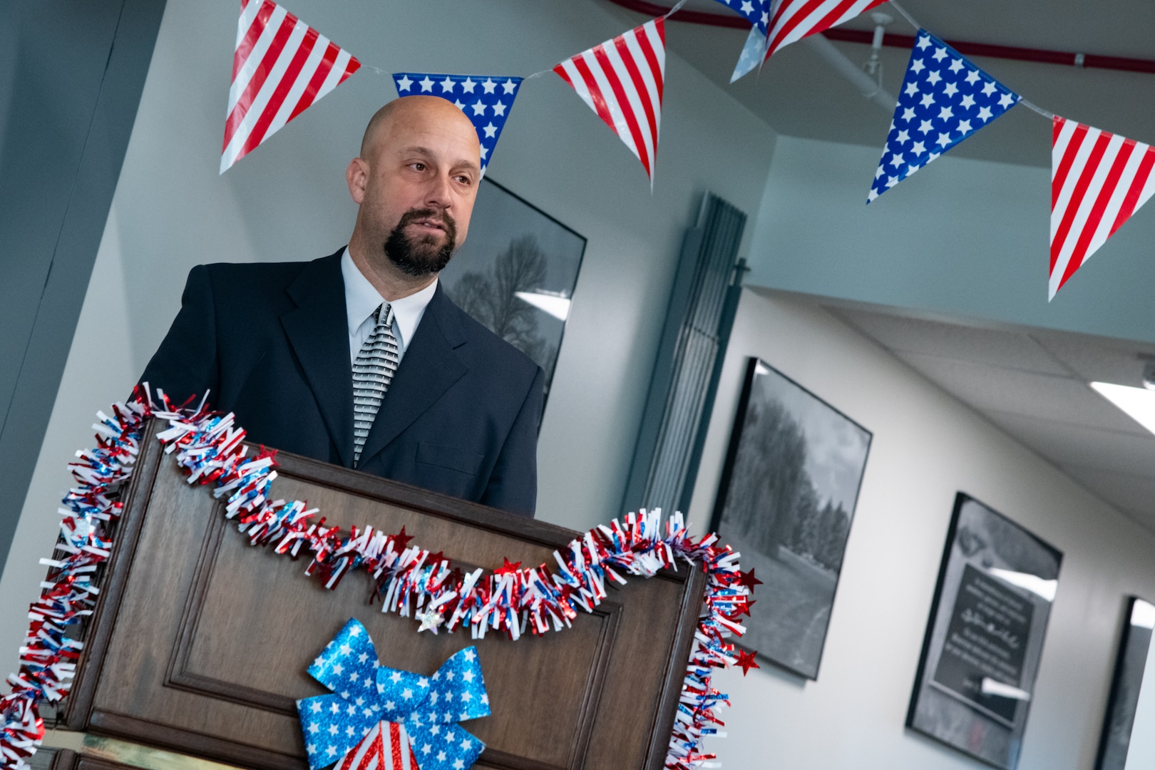 A man wearing a suit stands indoors behind a lectern speaking. There are American flag pennants behind him and red white and blue garland on the lectern.