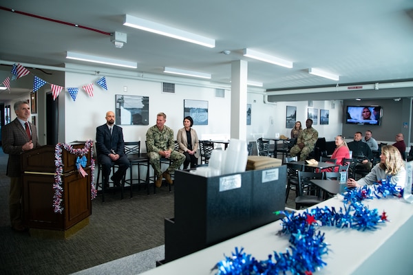 A man wearing a suit stands indoors behind a lectern speaking to a group of people seated at tables in the room. There are American flag pennants behind him and red white and blue garland on the lectern.