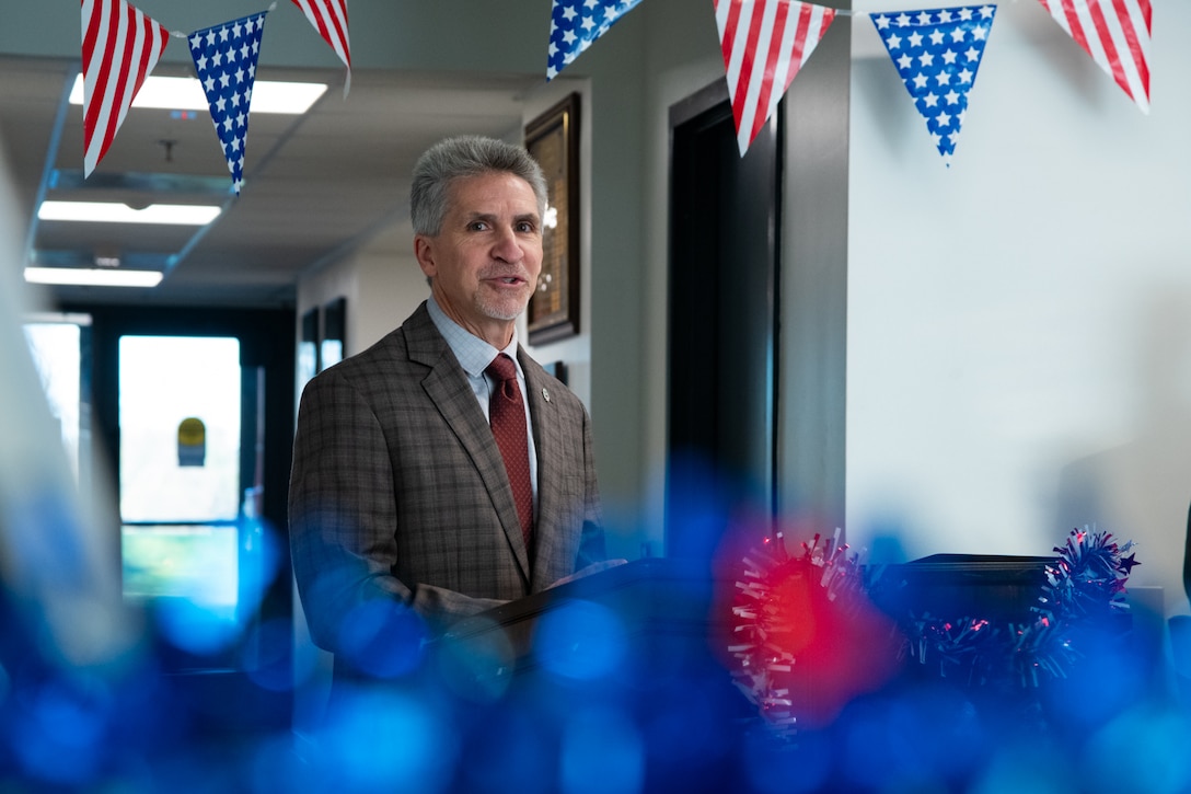 A man wearing a suit stands indoors behind a lectern speaking. There are American flag pennants behind him and red white and blue garland on the lectern.