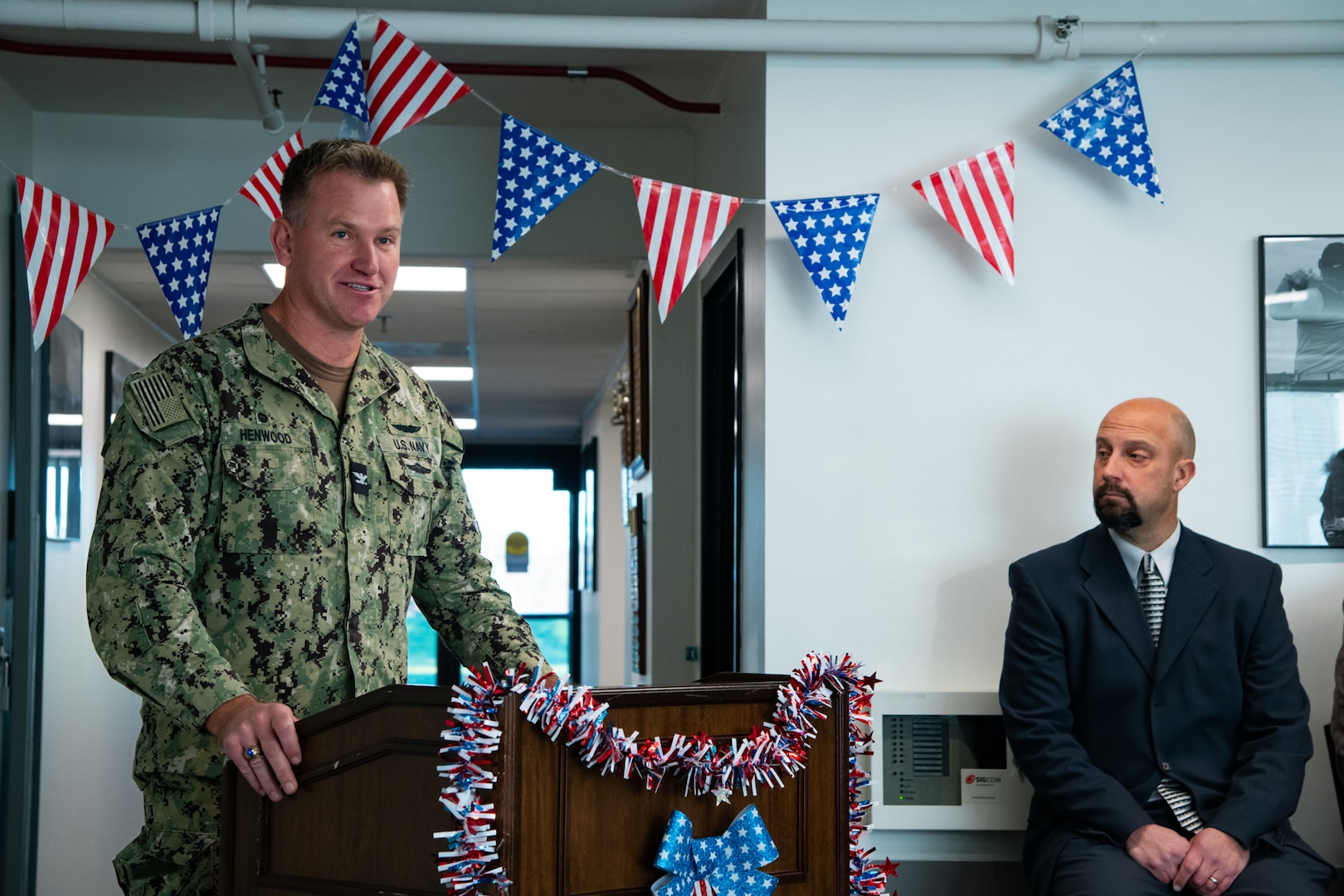 A man wearing a camouflage military uniform stands indoors behind a lectern speaking. There are American flag pennants behind him and red white and blue garland on the lectern.