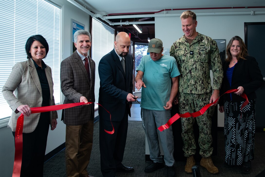 A group of six people stand inside behind a large red ribbon that has just been cut and is drifting toward the floor.