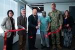 A group of six people stand inside behind a large red ribbon that has just been cut and is drifting toward the floor.