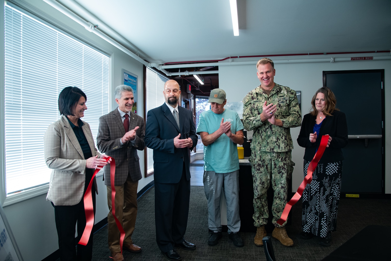 A group of six people stand inside behind a large red ribbon that has just been cut and is drifting toward the floor.