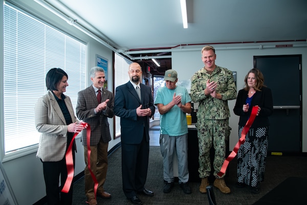 A group of six people stand inside behind a large red ribbon that has just been cut and is drifting toward the floor.