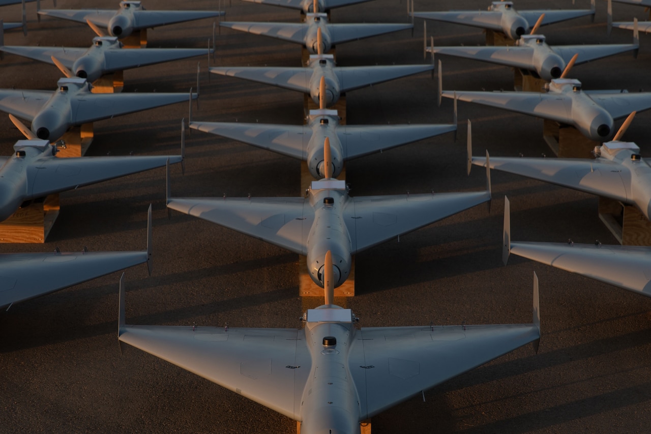 Combat attack drones are shown sitting on wood blocks in rows on a tarmac.