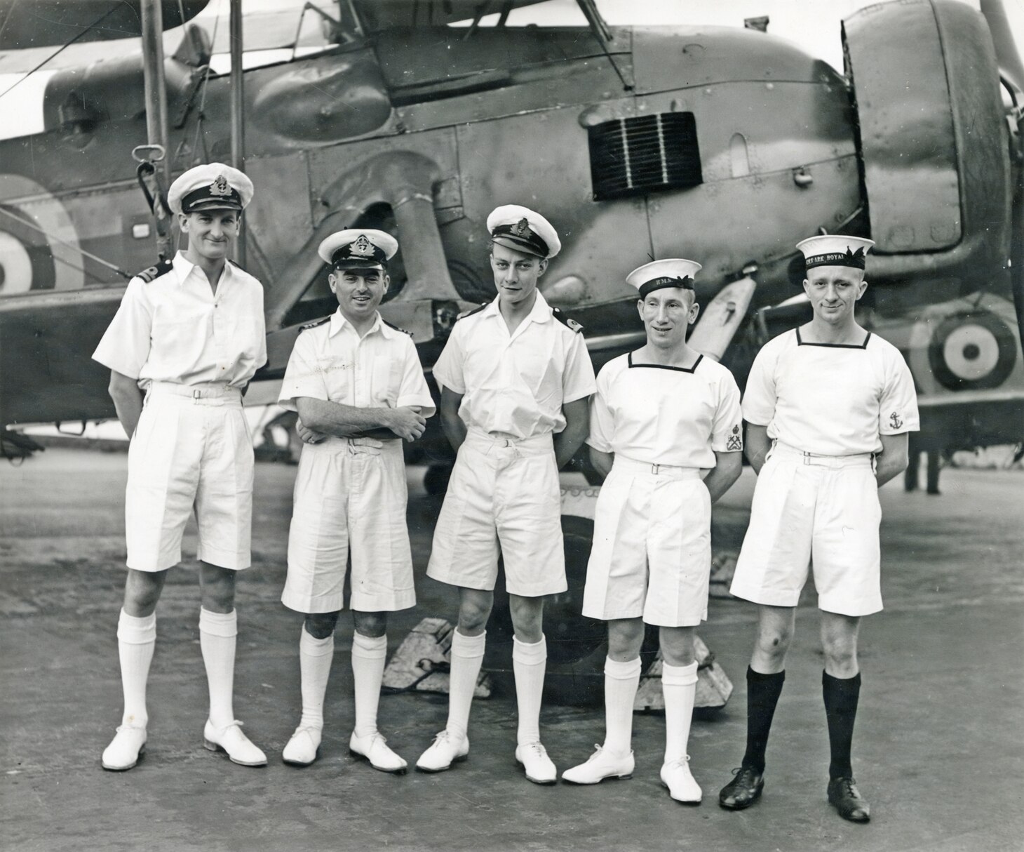 On the flight deck of the British aircraft carrier HMS Victorious, Lt. Cmdr. Eugene Esmonde, second from the left, stands with members of 825 Naval Air Squadron after receiving decorations received for their role in the sinking of the German battleship Bismark in May 1941. The officers received the Distinguished Service Cross, while Esmonde, the squadron commanding officer, received the Distinguished Service Order. The enlisted men received the Distinguished Service Medal. They are wearing what the U.S. Navy might call summer white uniforms. However, while most of the men are wearing white socks, the man, an enlisted air gunner, on the extreme right is obviously wearing black socks. Apparently, Royal Navy rules allow for a choice, and as this enlisted man was probably called away from his specific place to pose for the photo, his choice of footwear was slightly different.