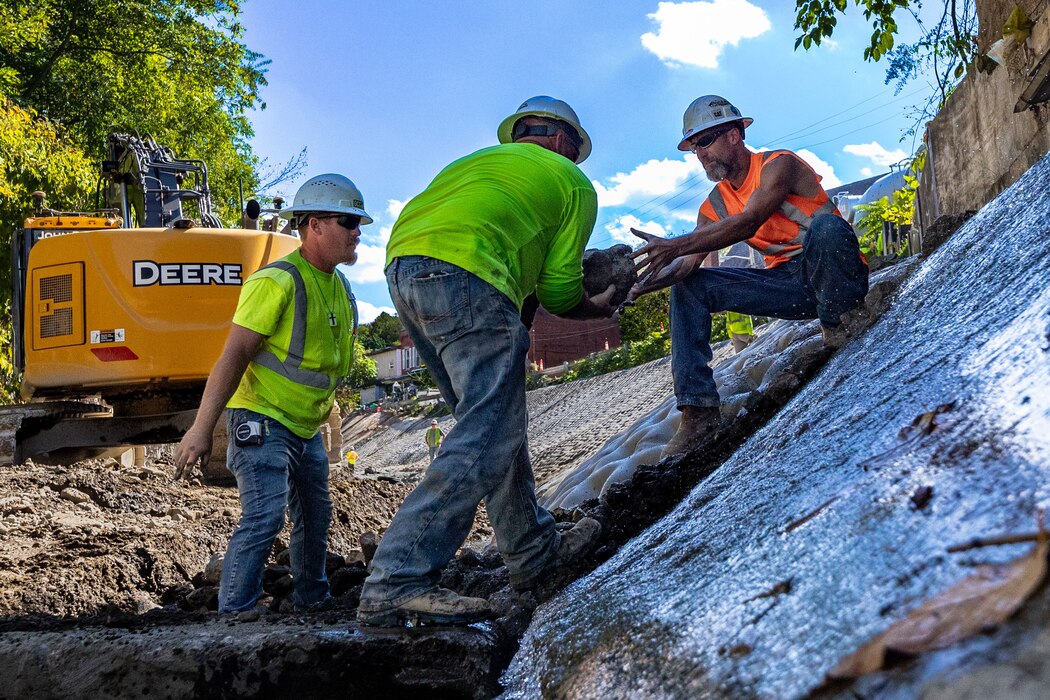 In a flood protection-project channel, a construction worker passes a stone to another construction worker.