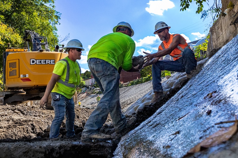 In a flood protection-project channel, a construction worker passes a stone to another construction worker.