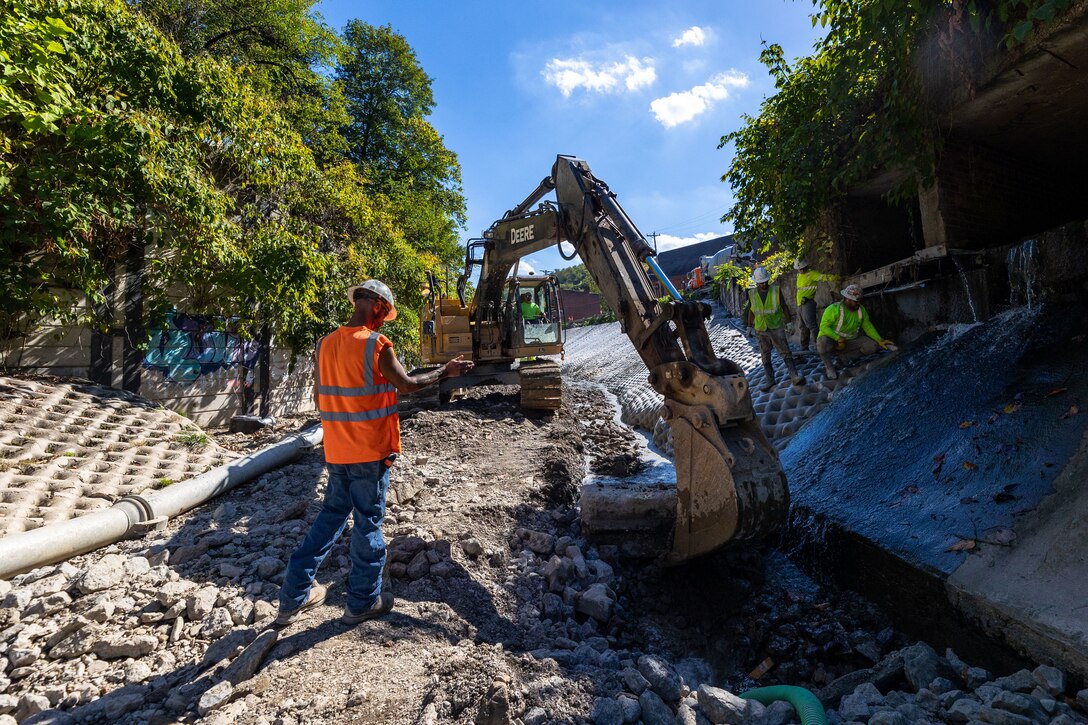 In a flood protection-project channel, construction workers stand near an excavator.