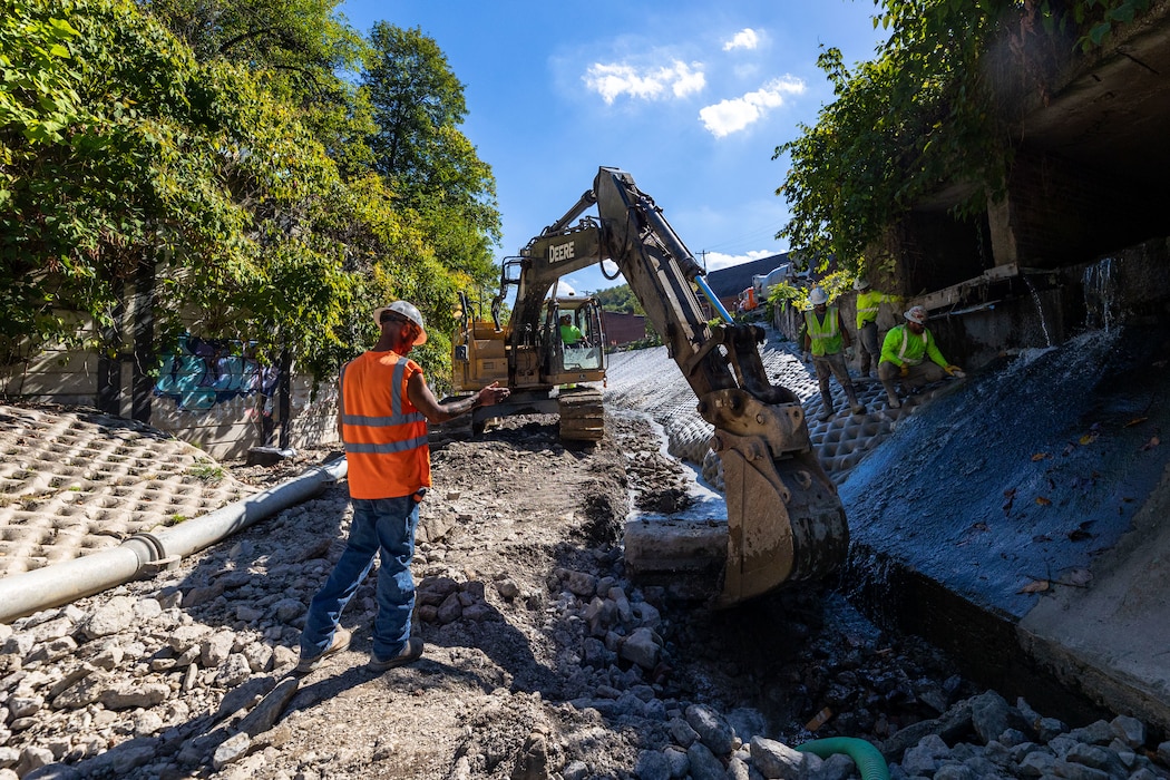 In a flood protection-project channel, construction workers stand near an excavator.