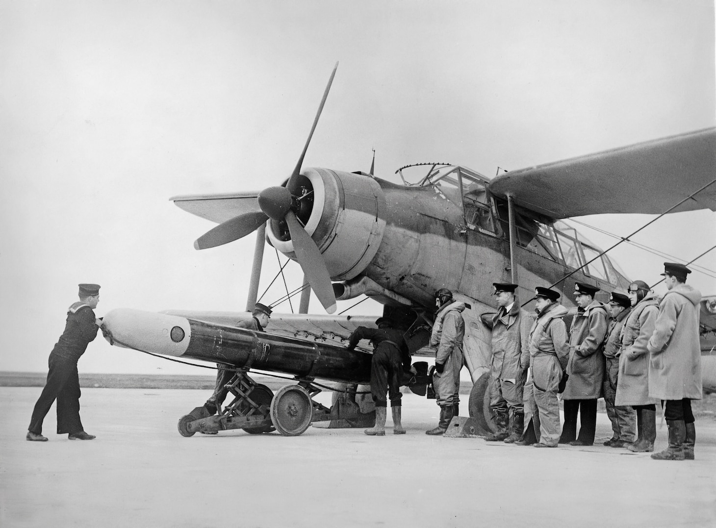 During a training operation, Albacore crews watch as a torpedo is loaded on one of the aircraft. The Albacore’s enclosed cockpit—as opposed to that of the Swordfish’s open flight stations—is shown.
