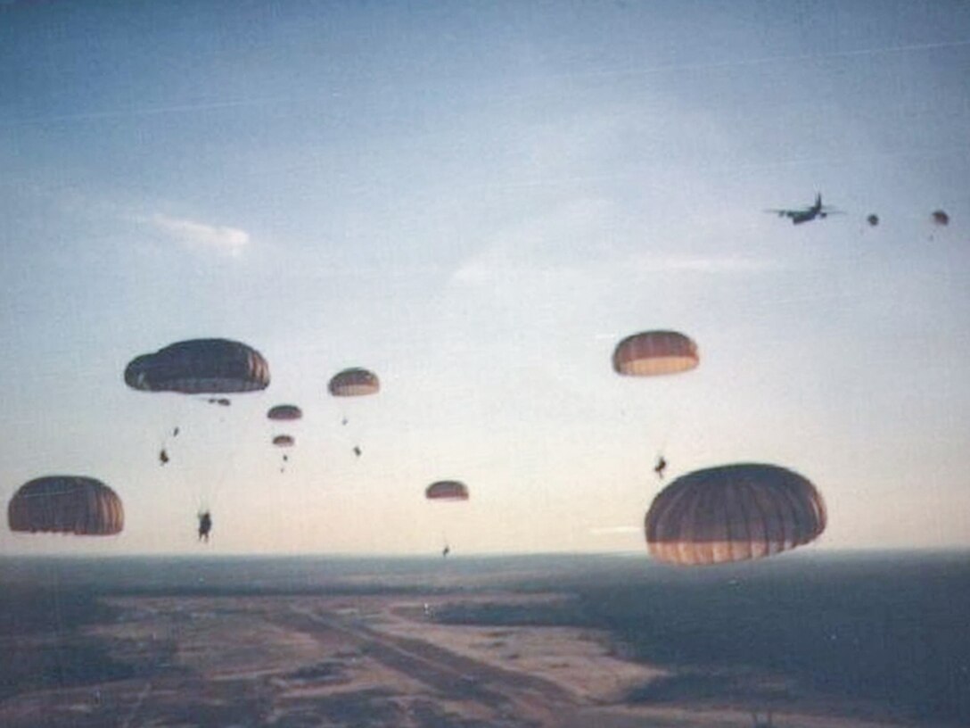 Soldiers descend with deployed Parachutes. Looks as if the photographer was also parachuting when the photo was taken,