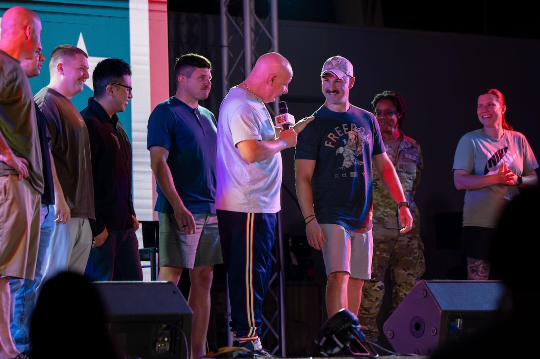 A person in casual attire and holding a microphone addresses a service member, also in casual attire, as other service members stand around them on a stage bathed in light.