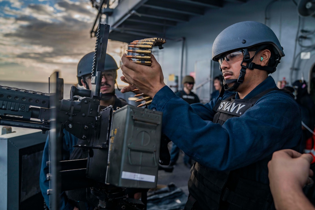 A sailor standing on the deck of a ship reloads a mounted machine gun with a belt of large bullets during daytime, with other sailors looking on.